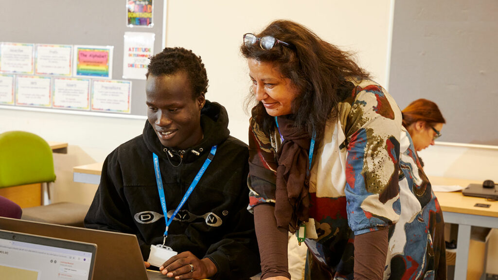 A student sat in front of a laptop with a tutor stood next to them