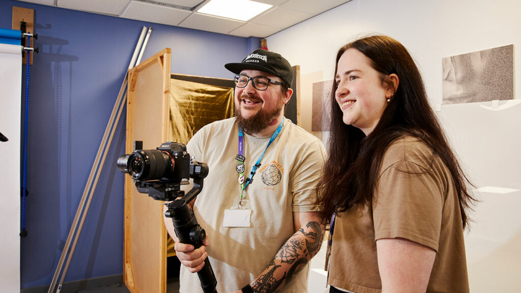 A student stood with their lecturer both looking at a camera