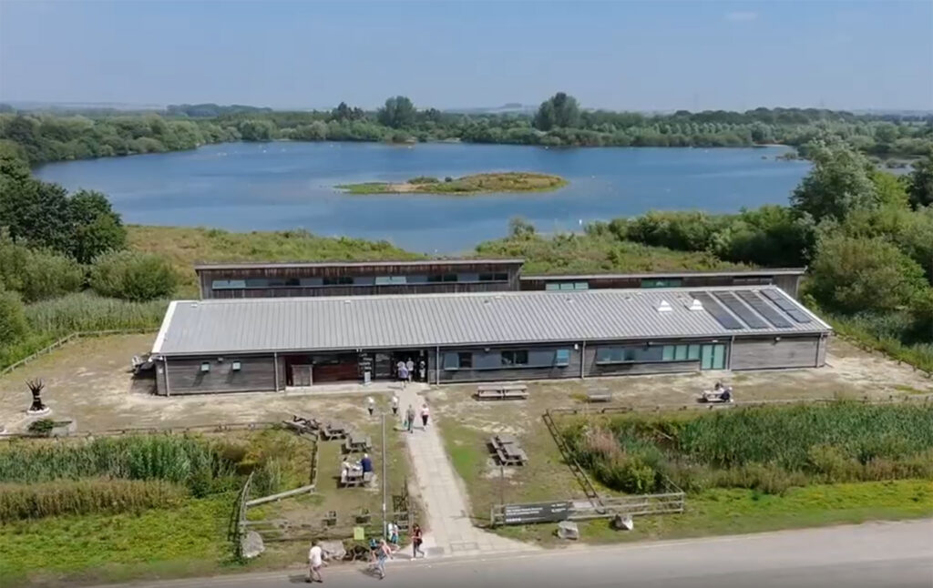 An aerial view of a long, one floor level wooden building, a path leading up to it and a large lake at the other side of it