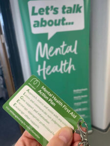 Mental Health First Aid green lanyard with a 'Lets talk about mental health' banner in the background