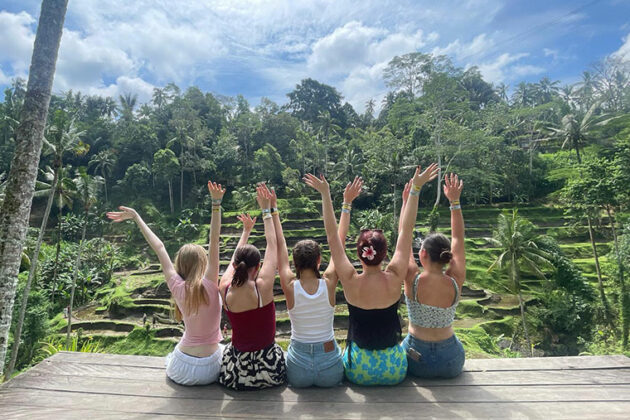 Five females sat on the edge of decking with their arms up in the air overlooking a beautiful view of trees and blue skies