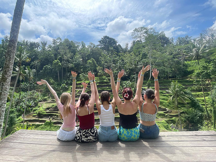 Five females sat on the edge of decking with their arms up in the air overlooking a beautiful view of trees and blue skies