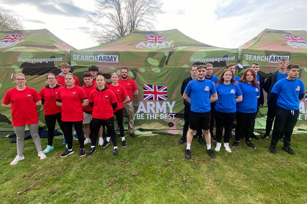 Two groups of student stood next to an army stand