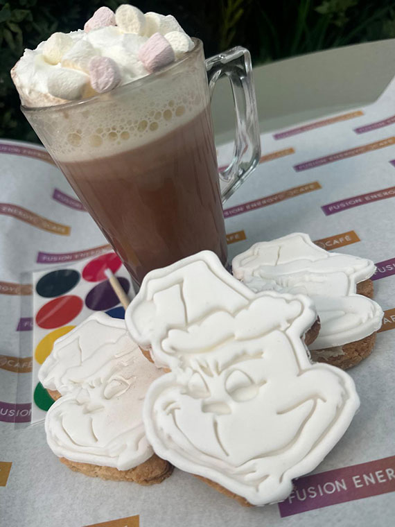 A hot chocolate topped with marshmallows and three Christmas cookies next to a paint palette ready to be painted