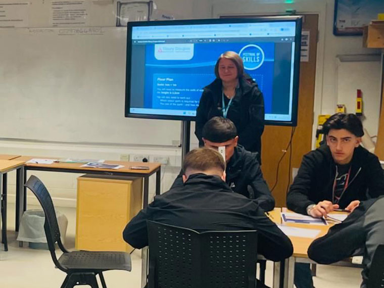 Four students sat around a desk in a classroom, tutor stood at the front