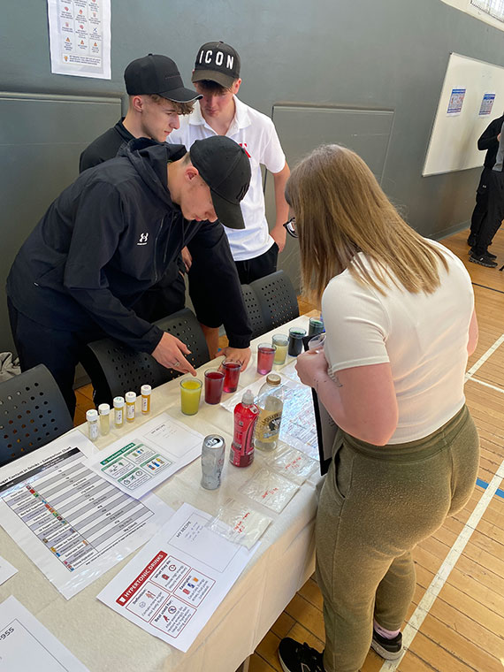 Three people stood behind a desk, one person stood infront of the desk looking at the different sports drinks