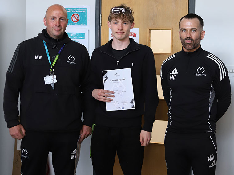 Momentous Football Academy awards winner holding a certificate stood with two members of staff