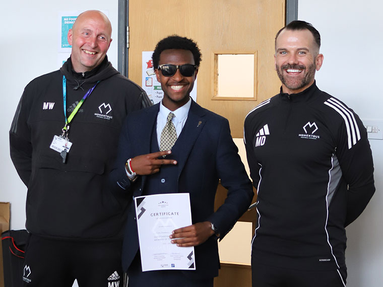Momentous Football Academy awards winner holding a certificate stood with two members of staff