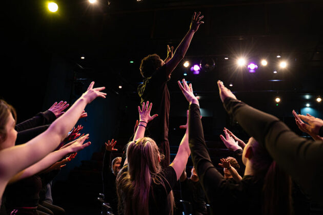 A group of students taking part in an acting performance sat on chairs with their hands in the air