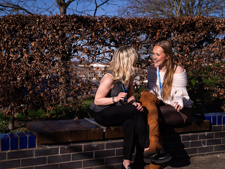 Two smiling students chatting sat on a wall with bushes behind them