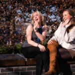 Two students sat on a wall smiling and waving