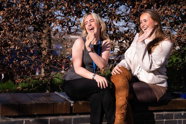 Two students sat on a wall smiling and waving