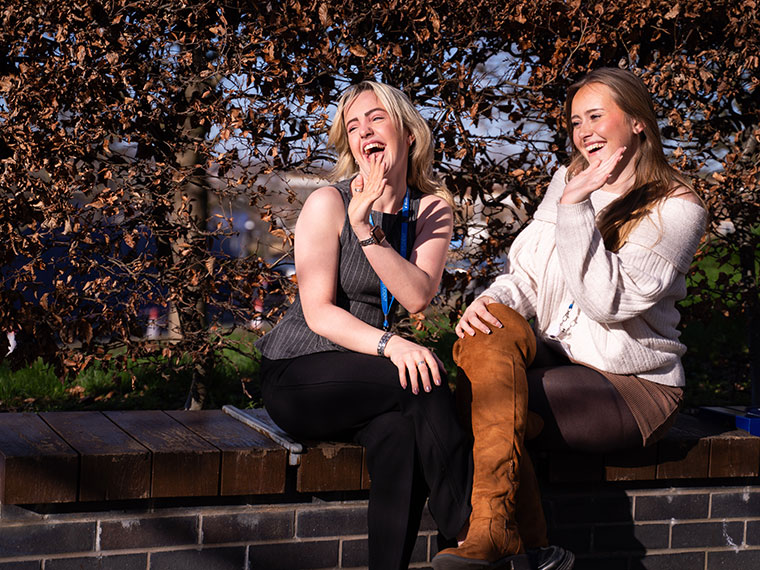 Two students sat on a wall smiling and waving