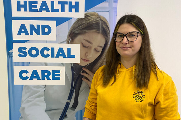 A female in a yellow long sleeved top stood in front of a banner that says 'Health and Social Care'