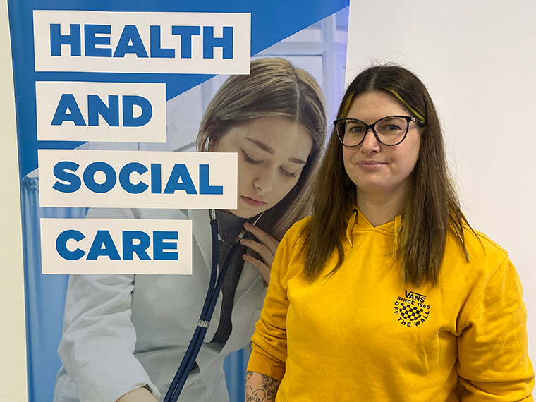 A female in a yellow long sleeved top stood in front of a banner that says 'Health and Social Care'