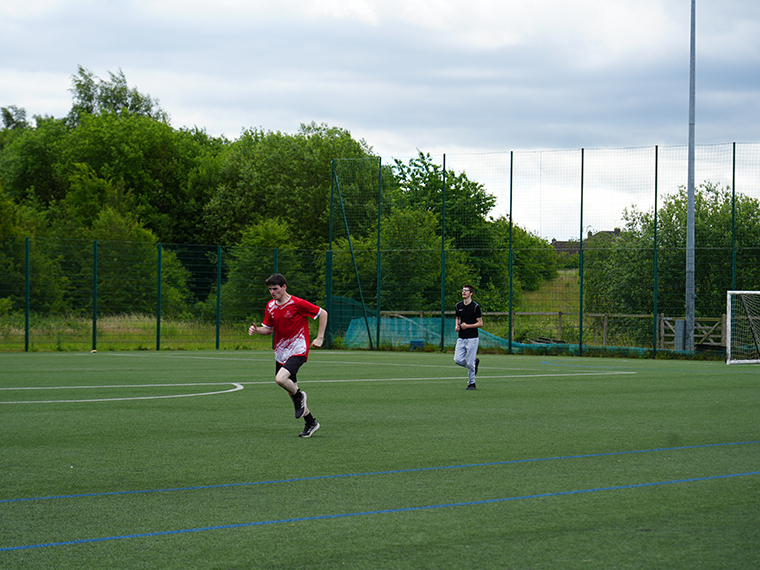 Someone in a red top and black shorts running across a football pitch, another person walking in the background