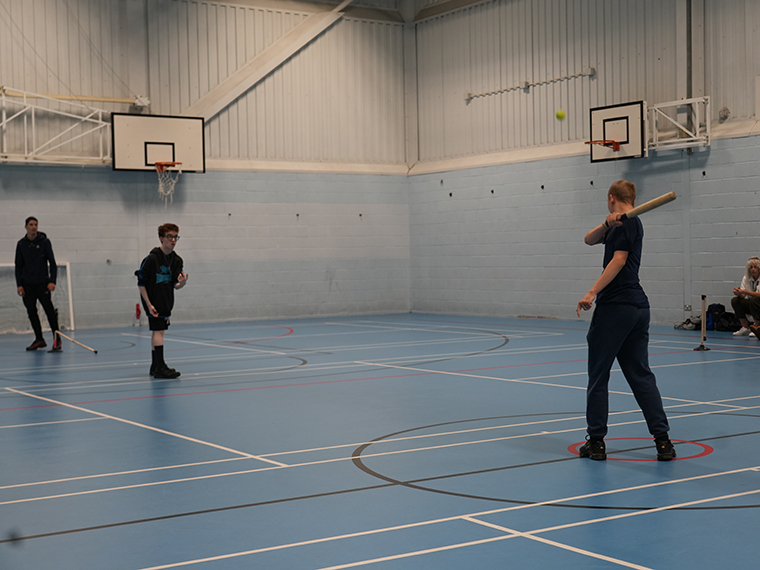 Three people stood in a gym, one person has just swung a baseball bat to hit a ball