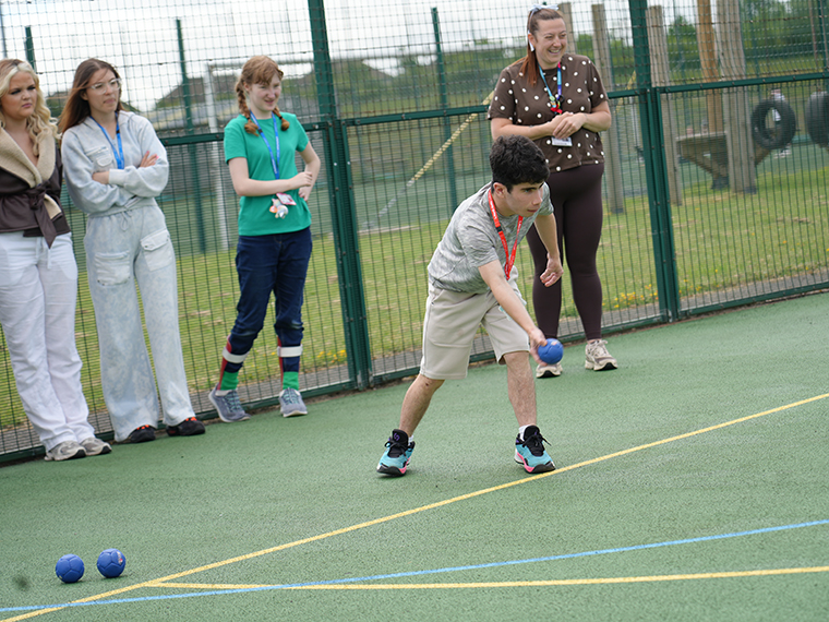 Someone wearing beige shorts and a white top about to throw a small blue bowling ball, four people stood behind watcching