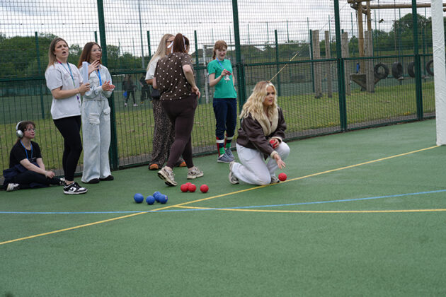 A female wearing black top and white trousers crouched down about to throw a small bowlilng ball, a small croud of people watching behind her