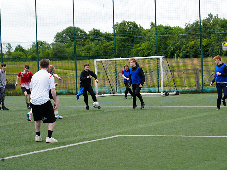 A group of people playing football on an outdoor pitch, a goal net is in the background