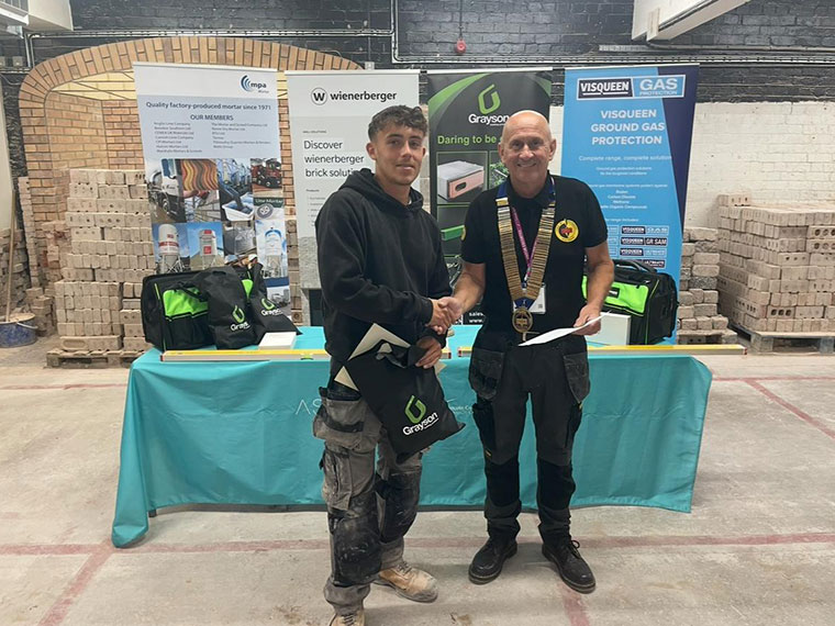 Two people stood shaking hands, one of them holding a certificate, with a table and banners in the background