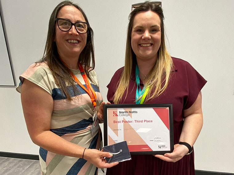 Two females, one wearing a stripey dress the other wearing a maroon dress, stood together holding a certificate between them