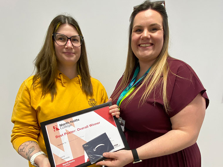 Two females, one wearing a yellow jumper the other wearing a maroon dress, stood together holding a certificate between them