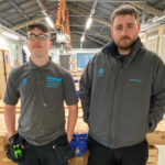 Two people stood in a joinery workshop, one wearing a grey tee shirt, the other wearing a grey fleece zipped up
