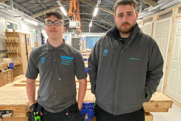 Two people stood in a joinery workshop, one wearing a grey tee shirt, the other wearing a grey fleece zipped up