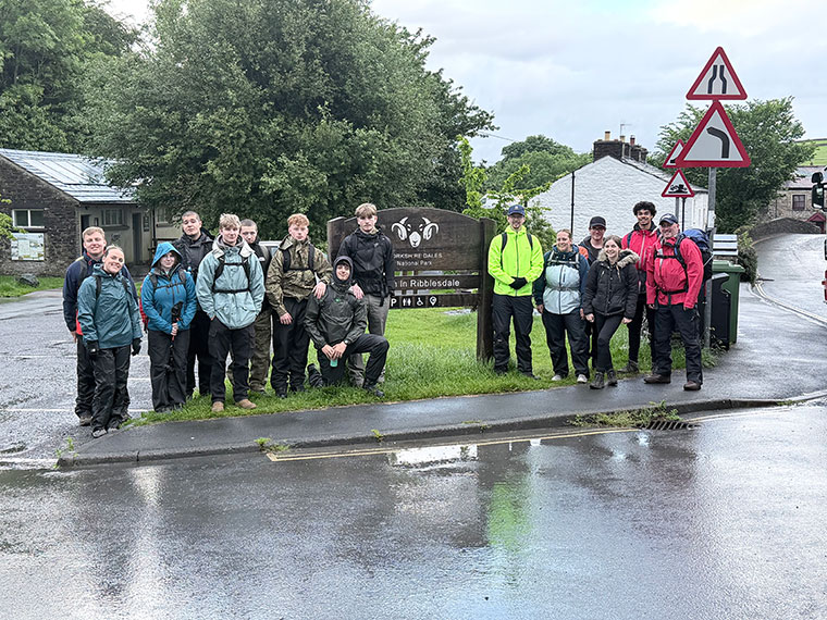 A group of people in walking gear stood in the rain next to a sign that says Ribblesdale