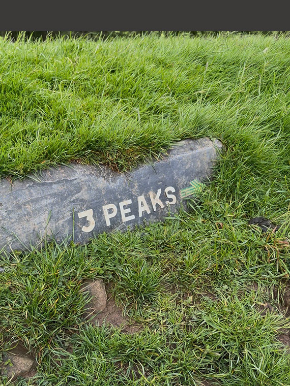 Green grass with a large flat stone showing with the words '3 Peaks' in white