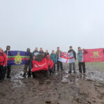 A group of people wearing walking gear stood on a muddy path, holding up four different flags including a red North Notts College flag.
