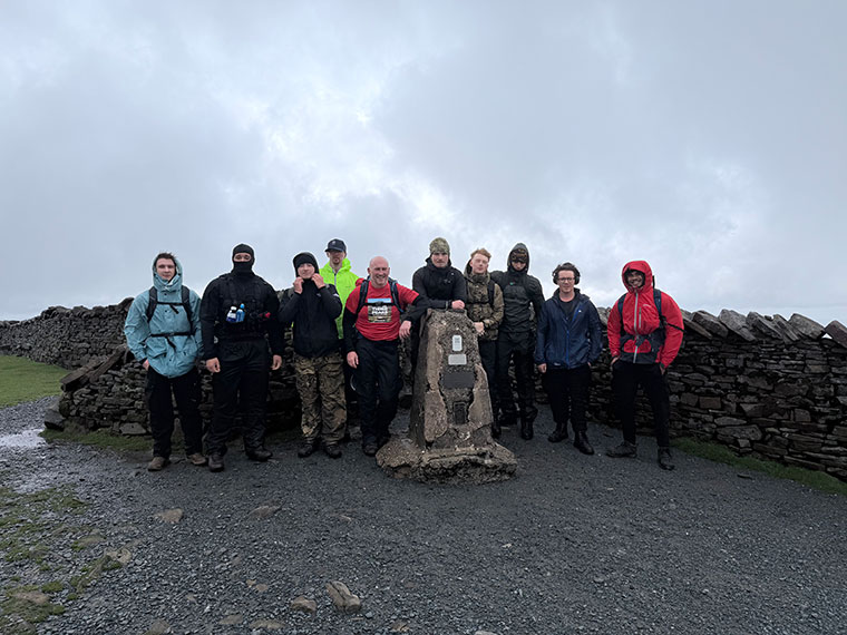 A group of people wearing walking gear stood next to a stone wall, the sky behind looking dark
