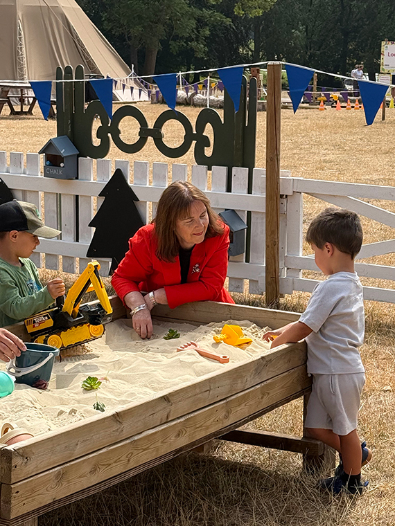 A female knelt down to a sandpit where two children are playing