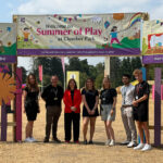 Seven people stood in a line with a banner above them reading 'Summer of Play'