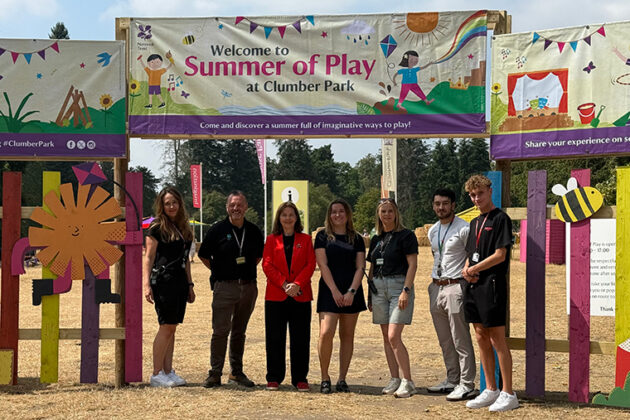 Seven people stood in a line with a banner above them reading 'Summer of Play'