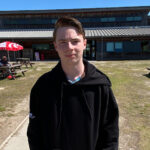 A male stood in front of a wooden building with grass in front of it and picnic tables