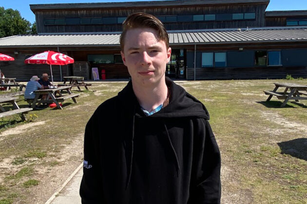A male stood in front of a wooden building with grass in front of it and picnic tables