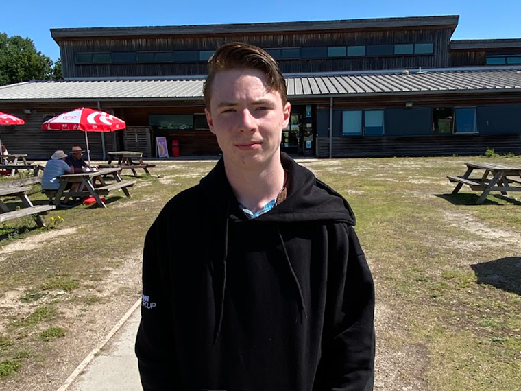 A male stood in front of a wooden building with grass in front of it and picnic tables