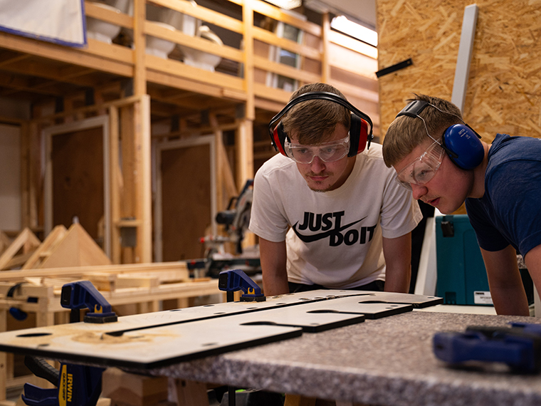 Two student wearing ear defenders bending down and looking at wood on a table, frames made out of wood in the background