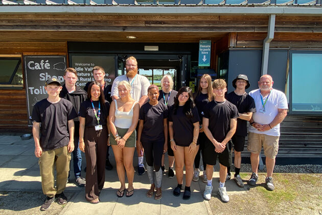 A group of people stood outside in the sun, a wooden building behind them