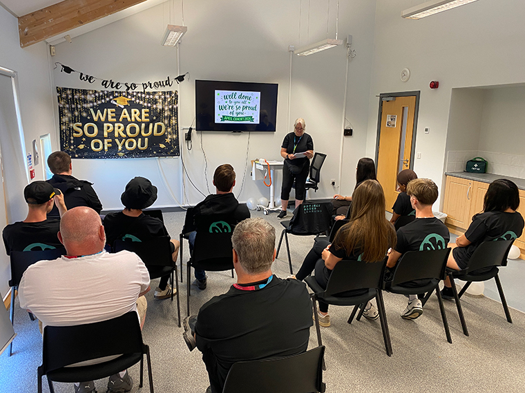 A group of people seated looking towards a screen on the wall and a person stood at the front presenting to them