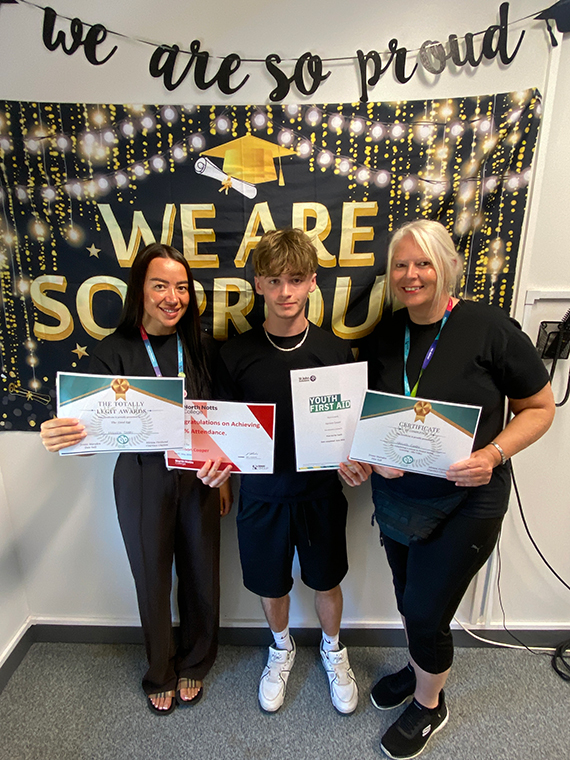 Two females stood either side of a male, holding four certificates, two banners behind them saying 'we are so proud'