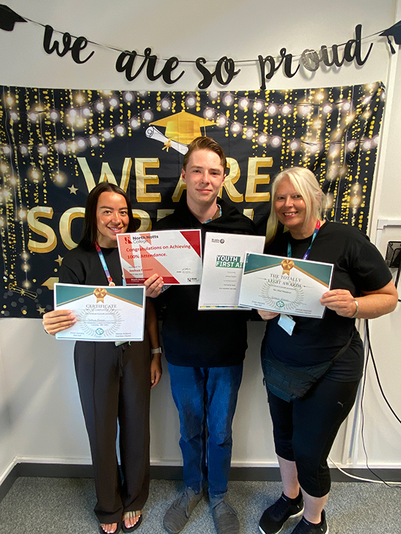 Two females stood either side of a male, holding four certificates, two banners behind them saying 'we are so proud'