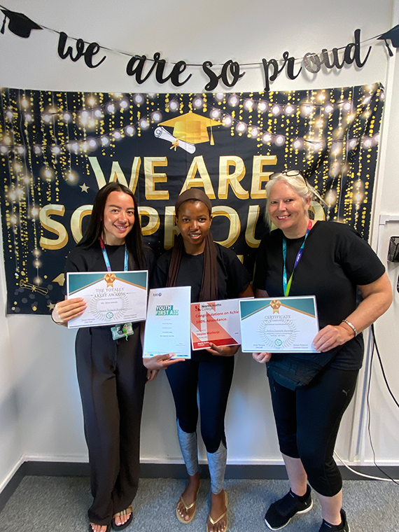 Three females stood together holding four certificates, two banners behind them saying 'we are so proud'