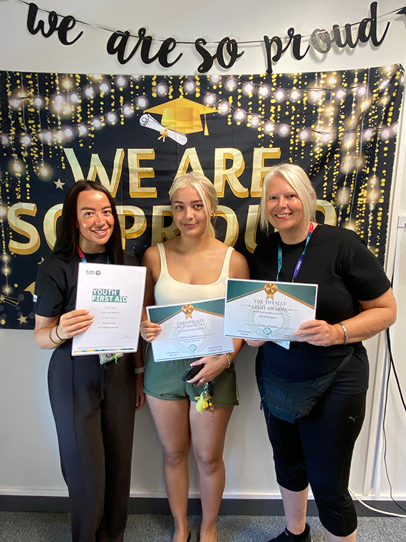 Three females stood together holding two certificates, two banners behind them saying 'we are so proud'