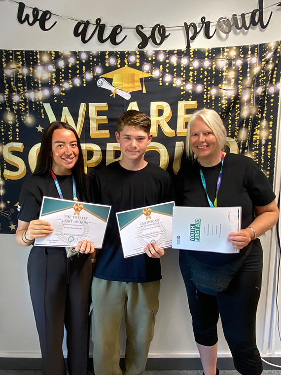 Two females stood either side of a male, holding three certificates, two banners behind them saying 'we are so proud'