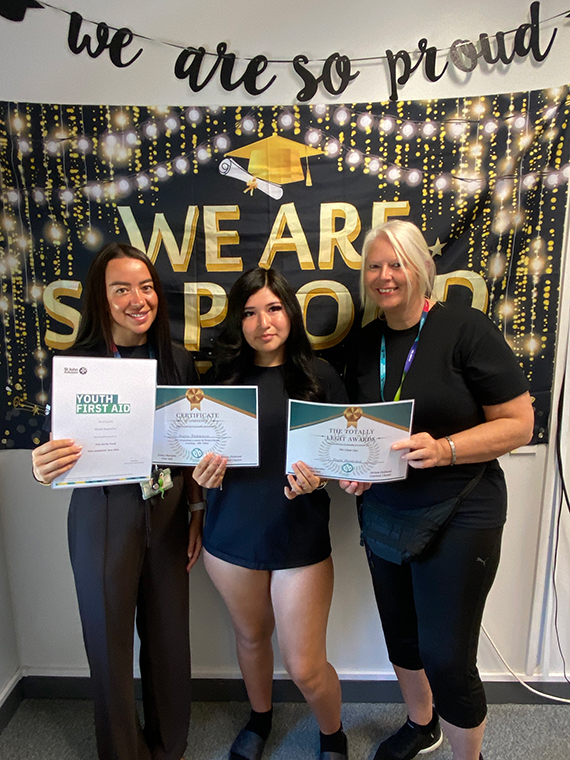 Three females stood together holding three certificates, two banners behind them saying 'we are so proud'