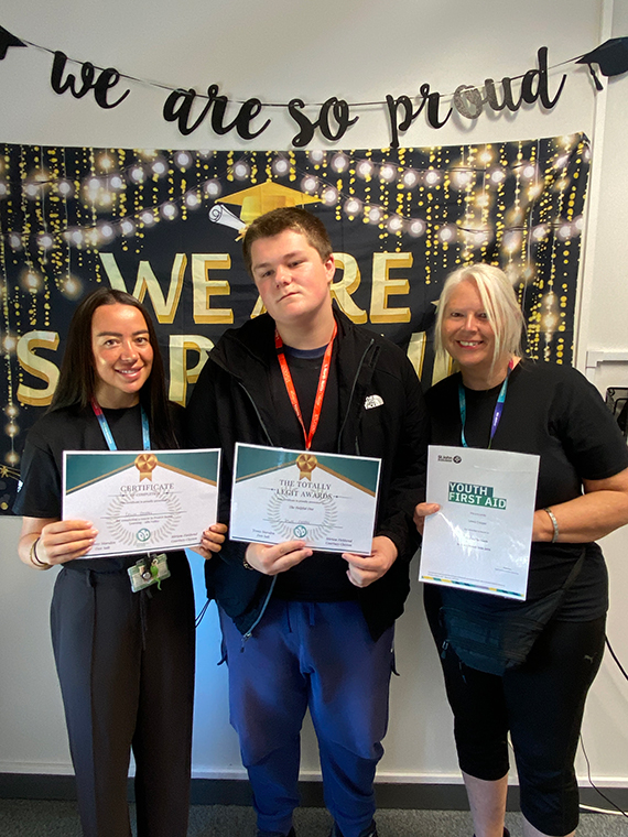 Two females stood either side of a male, holding three certificates, two banners behind them saying 'we are so proud'