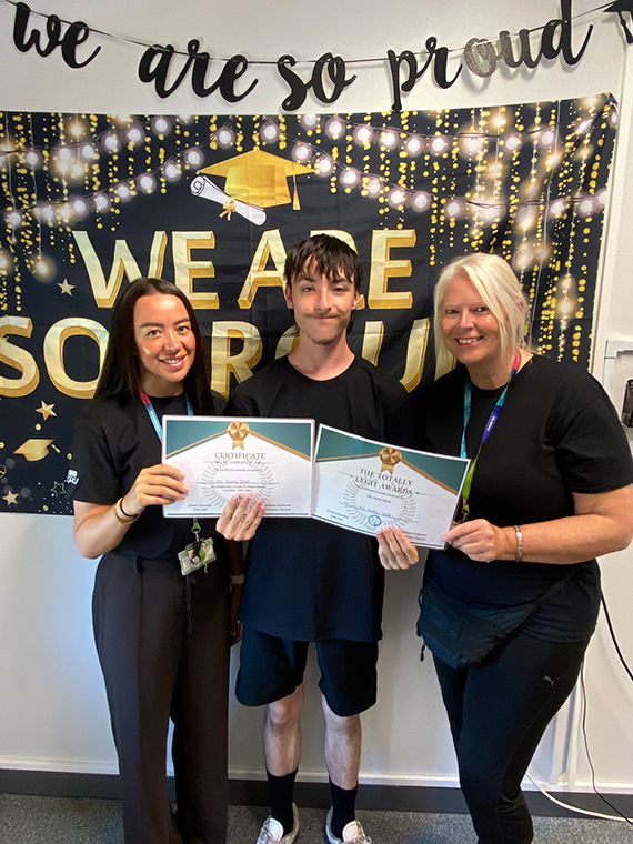 Two females stood either side of a male, holding two certificates, two banners behind them saying 'we are so proud'